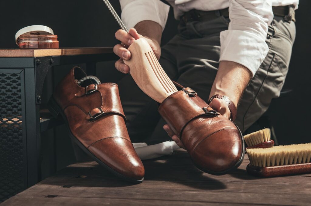 Muškarac umeće napinjače u kožne cipele. Pored njega se nalaze proizvodi za održavanje i njegu obuće.