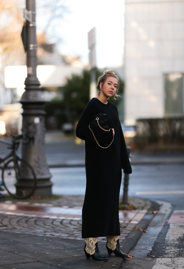 Djevojka u ženskim kaubojskim čizmama s dezenom u kombinaciji s crnom maxi haljinom stoji uz cestu.