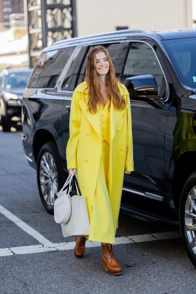 Model nosi smeđe čizme s debelim potplatom u kombinaciji s žutom haljinom i sivom torbicom.