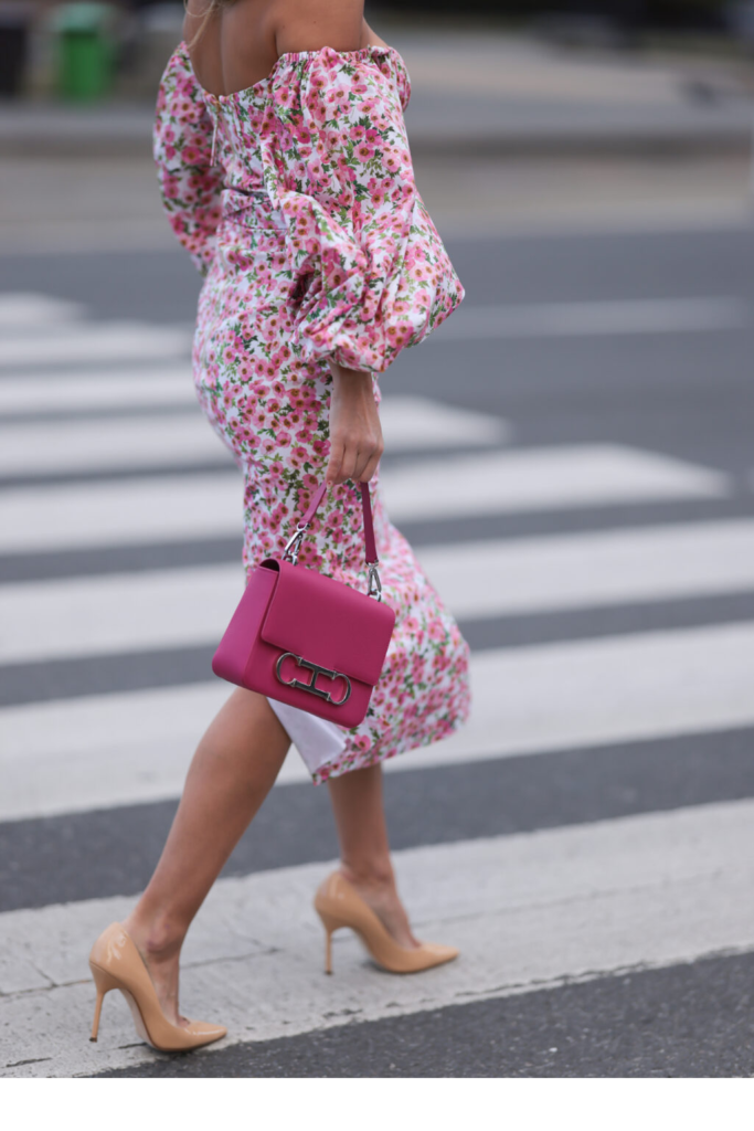Žena u cvjetnoj haljini s puf rukavima spuštenih ramena s žarko ružičastom torbicom i bež lakiranim visokim petama
